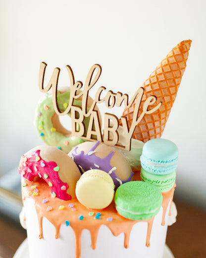 Wooden topper reads "Welcome Baby," surrounded by colorful sweets and a cone on a decorated cake.