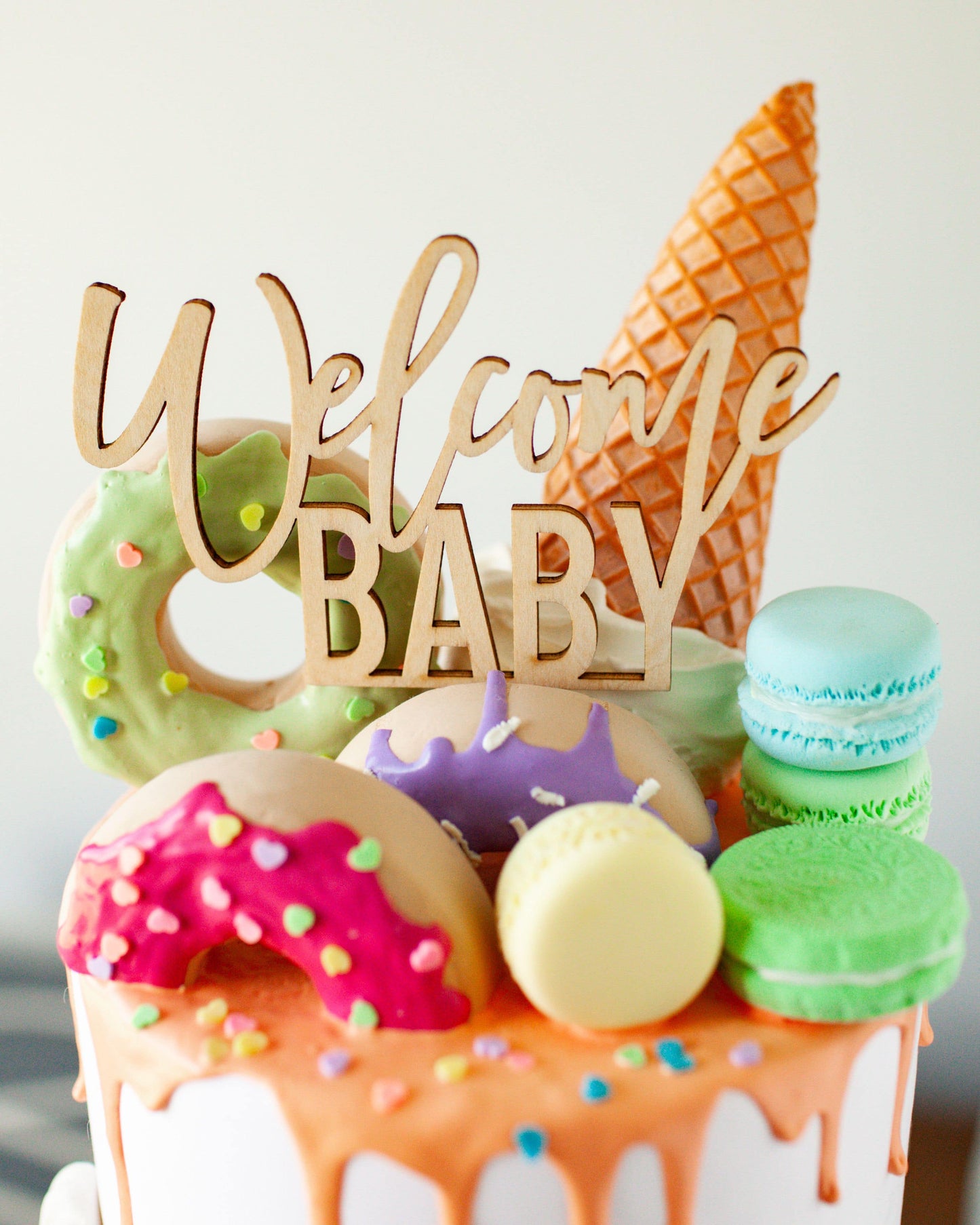 Wooden topper reads "Welcome Baby" above colorful sweets including donuts, macarons, and an ice cream cone.