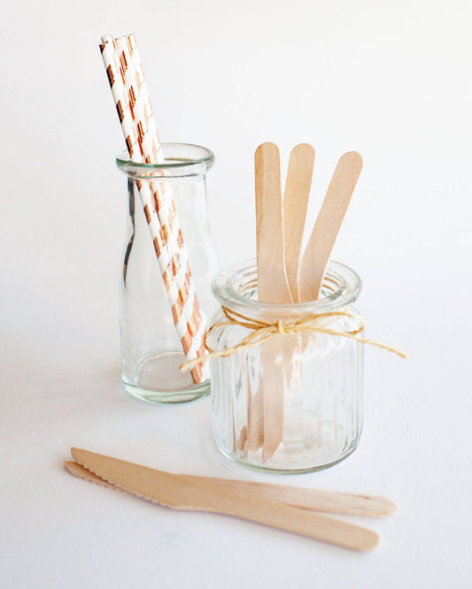 Two glass containers hold wooden utensils and striped straws, arranged on a light background.