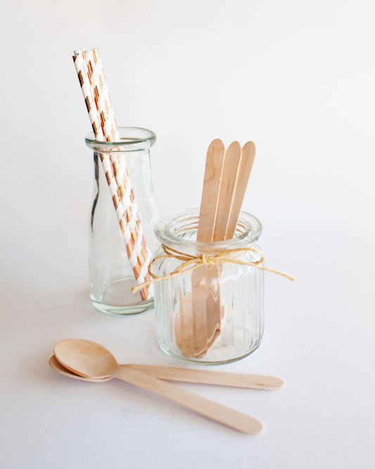 A clear glass jar holds wooden sticks, accompanied by wooden spoons and striped paper straws on a light surface.